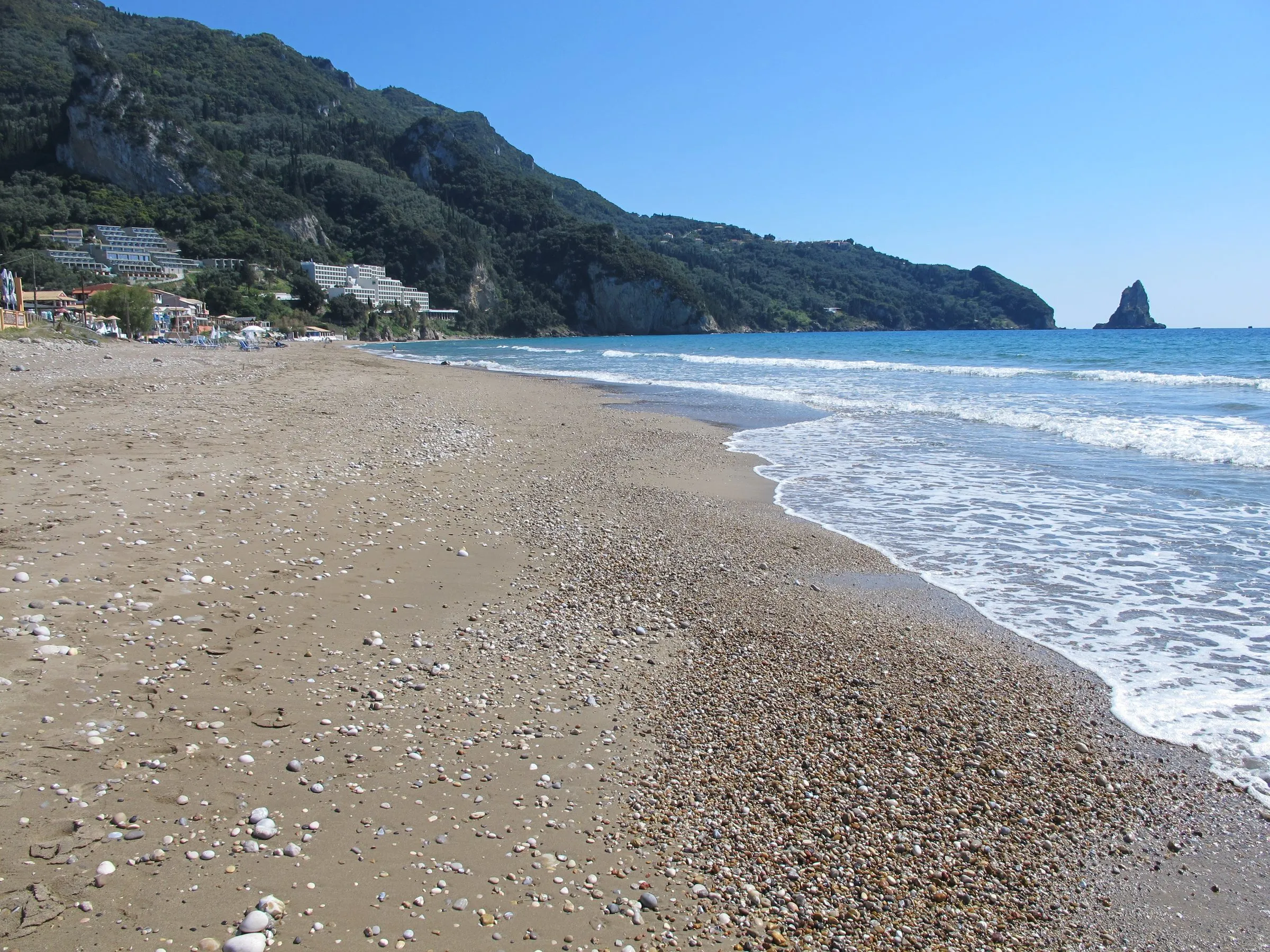 Kies-Sand-Strand von Agios Gordios mit dem Felsen Ortholithos im Meer